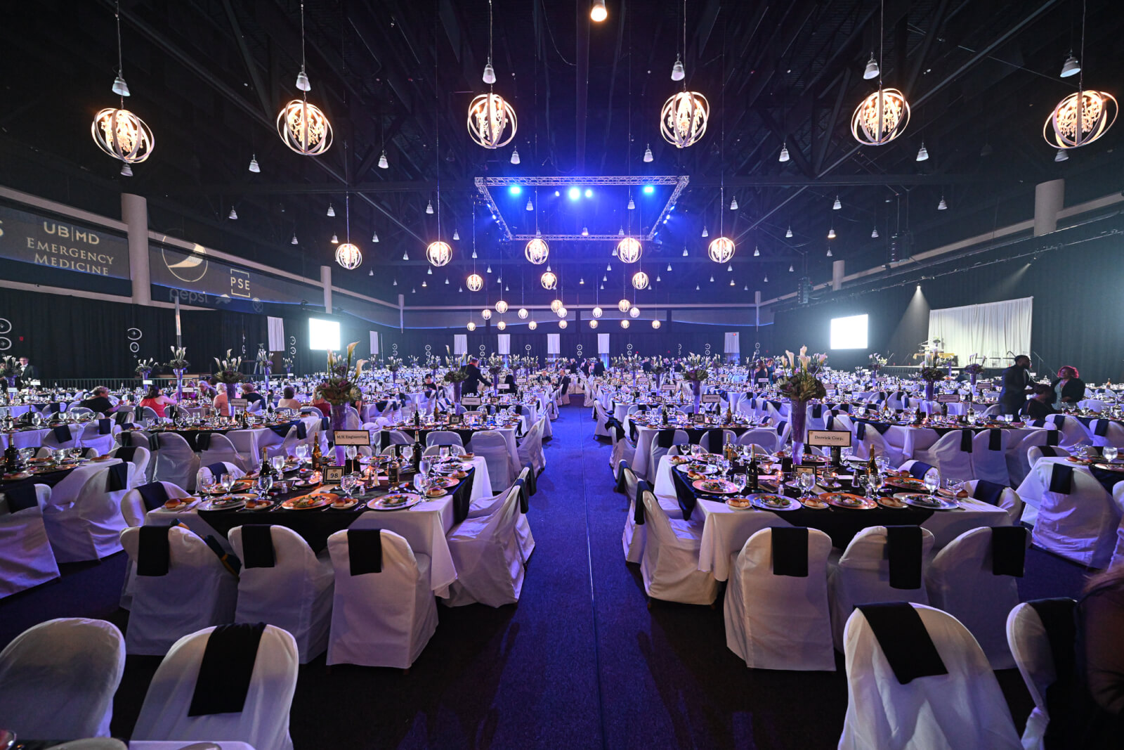 A large ballroom is set for a formal dinner, complete with while chair covers and hanging light medallions.